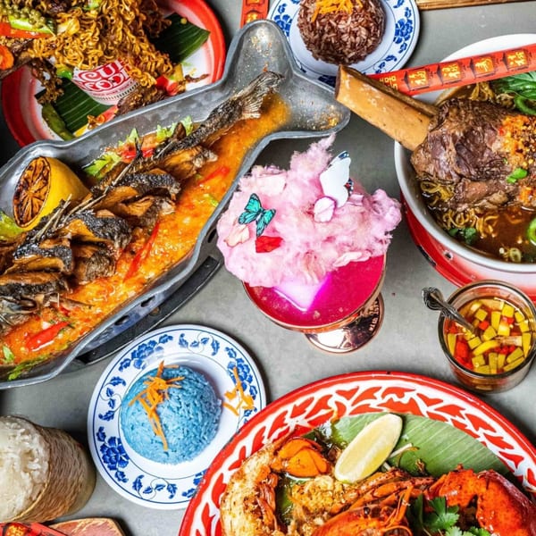 A vibrant spread of Thai dishes, including a whole fish, noodles, soup, blue rice, and a pink cocktail.