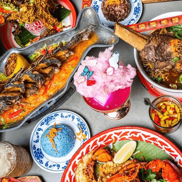 Overhead view of a vibrant Thai feast with whole fish, noodles, blue rice, lobster, and a pink drink.