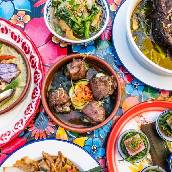 A vibrant overhead shot of various Thai dishes, including soups, beef skewers, and tuna, on a floral tablecloth.