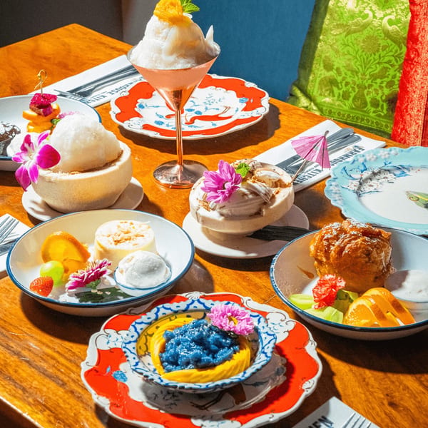A wooden table laden with various colorful Thai desserts, including coconut ice cream and mango sticky rice.