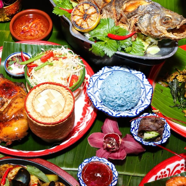 A vibrant Thai feast with fried fish, papaya salad, blue rice, grilled chicken, and various sauces on banana leaves.