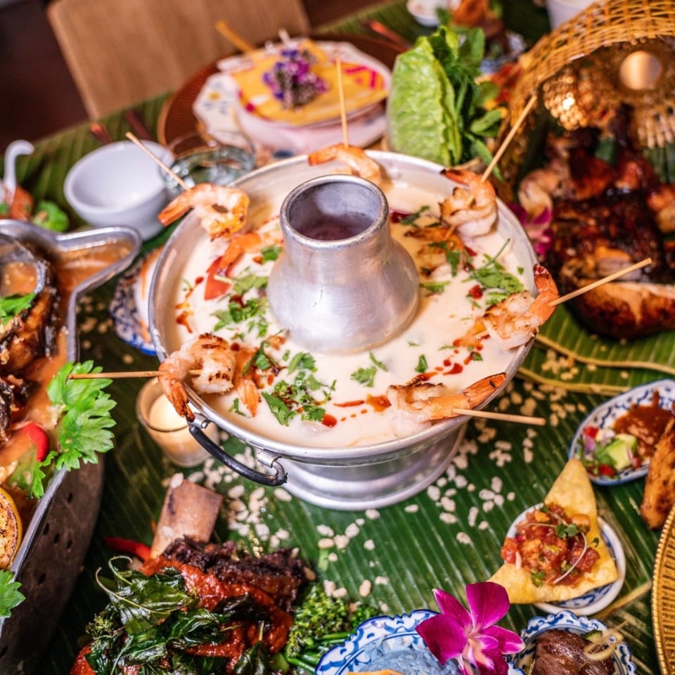 A Thai hot pot with shrimp skewers, surrounded by various Thai dishes on a banana leaf.