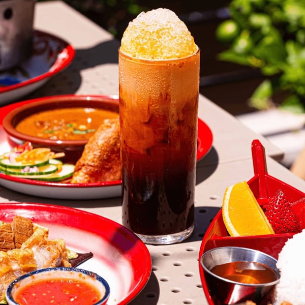 A tall glass of Thai iced tea with shaved ice, surrounded by various Thai dishes on red plates.