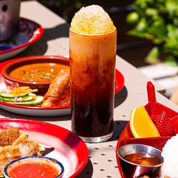 A tall glass of iced Thai tea with shaved ice, surrounded by various Thai dishes on red plates.